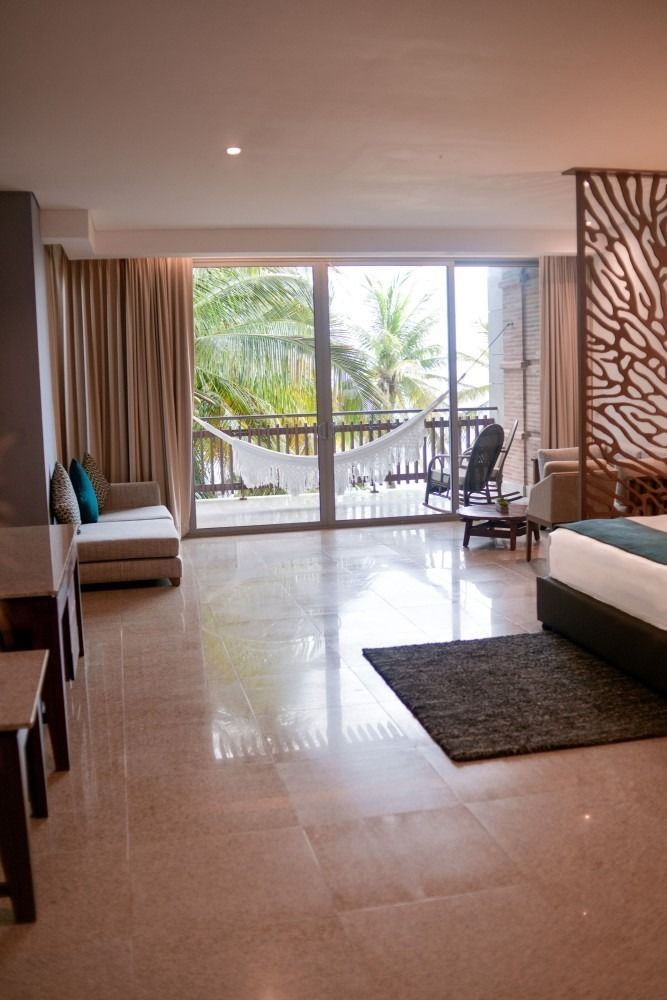 Bright hotel room with sliding glass doors, balcony with hammock, couch, rug, and decorative wooden panel.