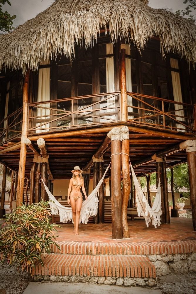 Woman in a bikini stands on a red-brick porch in front of a multi-story thatched-roof building.