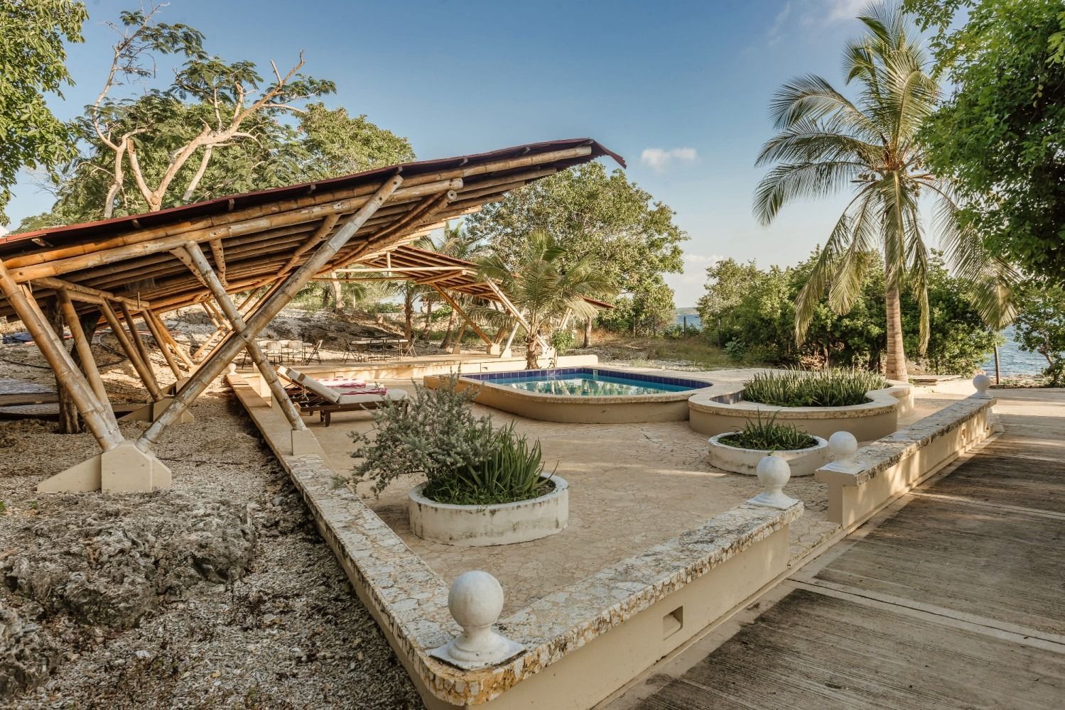 Pool area with shade structure, tropical plants, and a partial ocean view under a blue sky.
