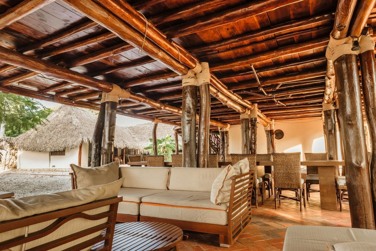 Covered outdoor seating area with wooden beams, furniture, and thatched-roof building in background.