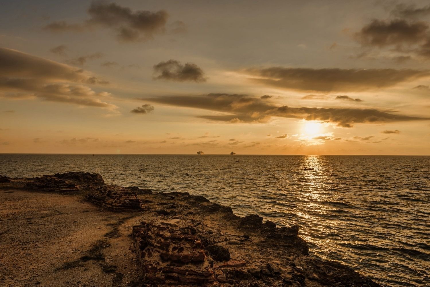 Sunset over ocean, golden light reflecting on water, rocky coastline in foreground.