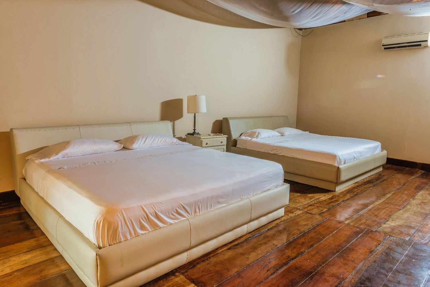 Two beds in a room with hardwood floors and beige walls; a bedside lamp and air conditioner are visible.