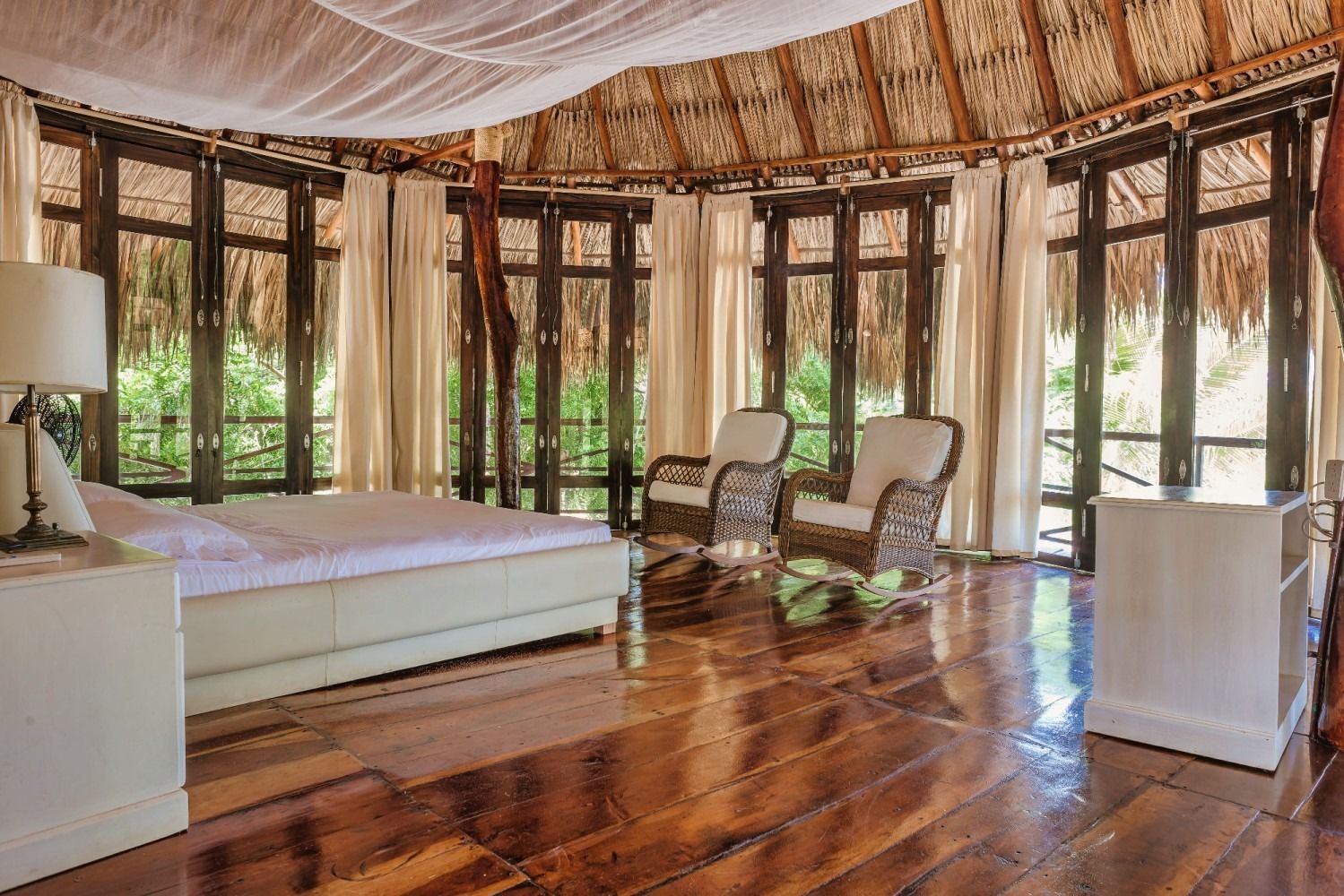 Bedroom with thatched roof, wooden floors, large windows, bed, two armchairs.