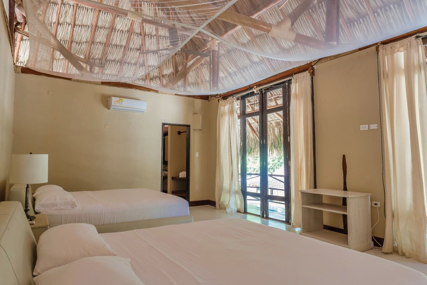 Bedroom with two beds, thatched roof, and large glass doors. Creamy walls, white linens, and simple decor.