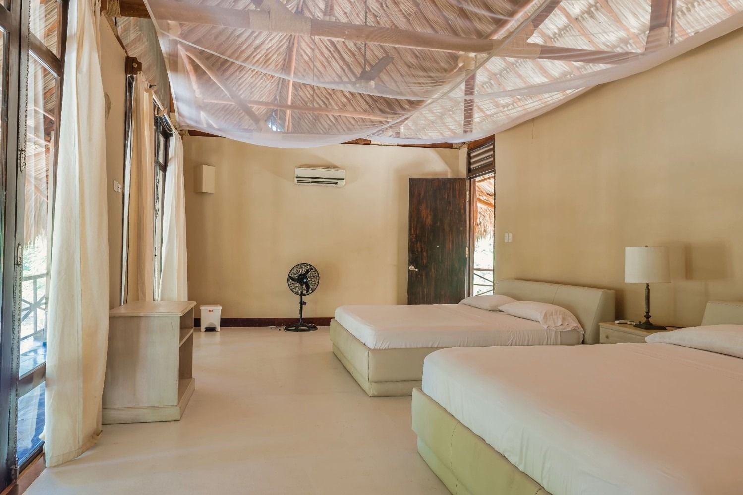 Bedroom with two beds, cream walls, open windows, fan, air conditioning, and a thatched ceiling.