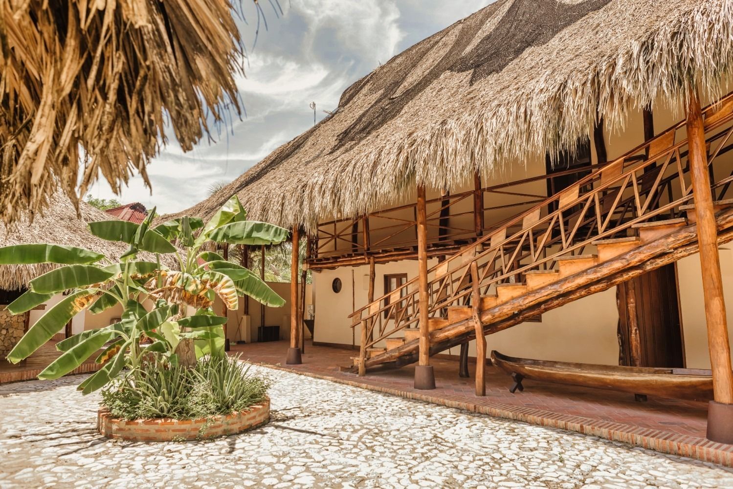 Courtyard with thatch roof buildings, wooden stairs, and tropical plants.