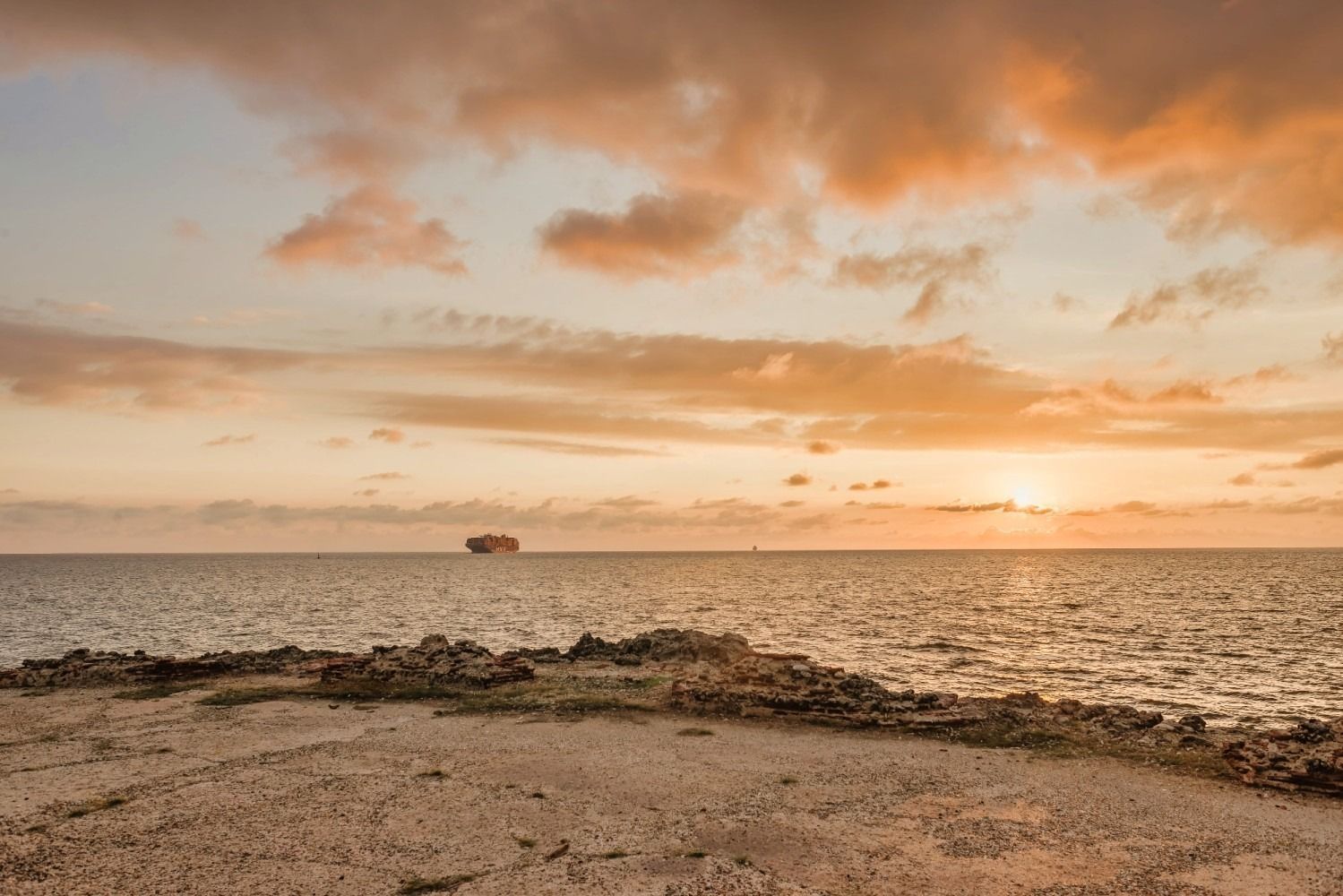 Ocean view at sunset, with a silhouette of a ship on the horizon, and rugged shoreline in foreground.