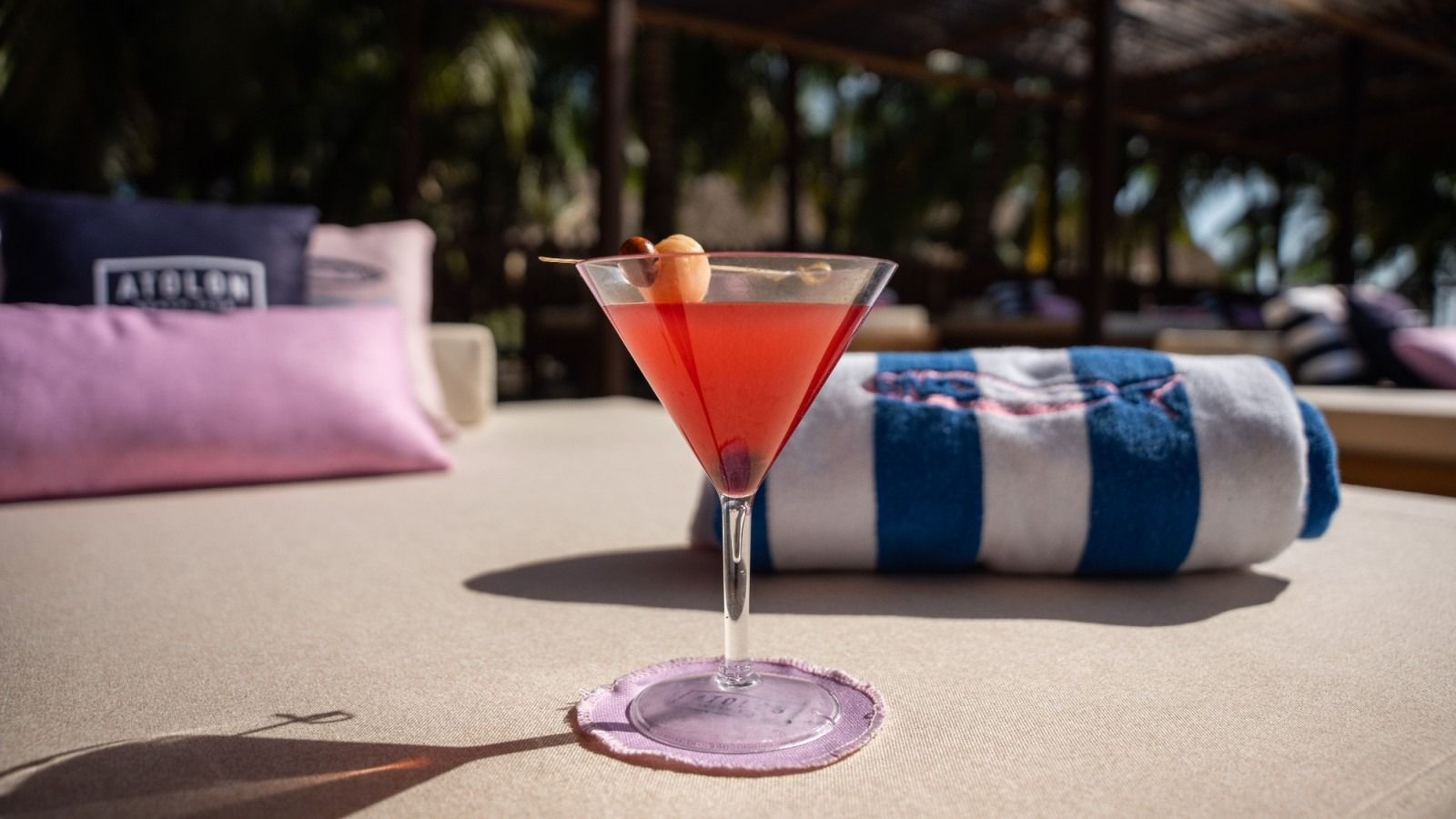 Cocktail in a martini glass on a lilac coaster, by a rolled towel and pillows on a lounge bed.