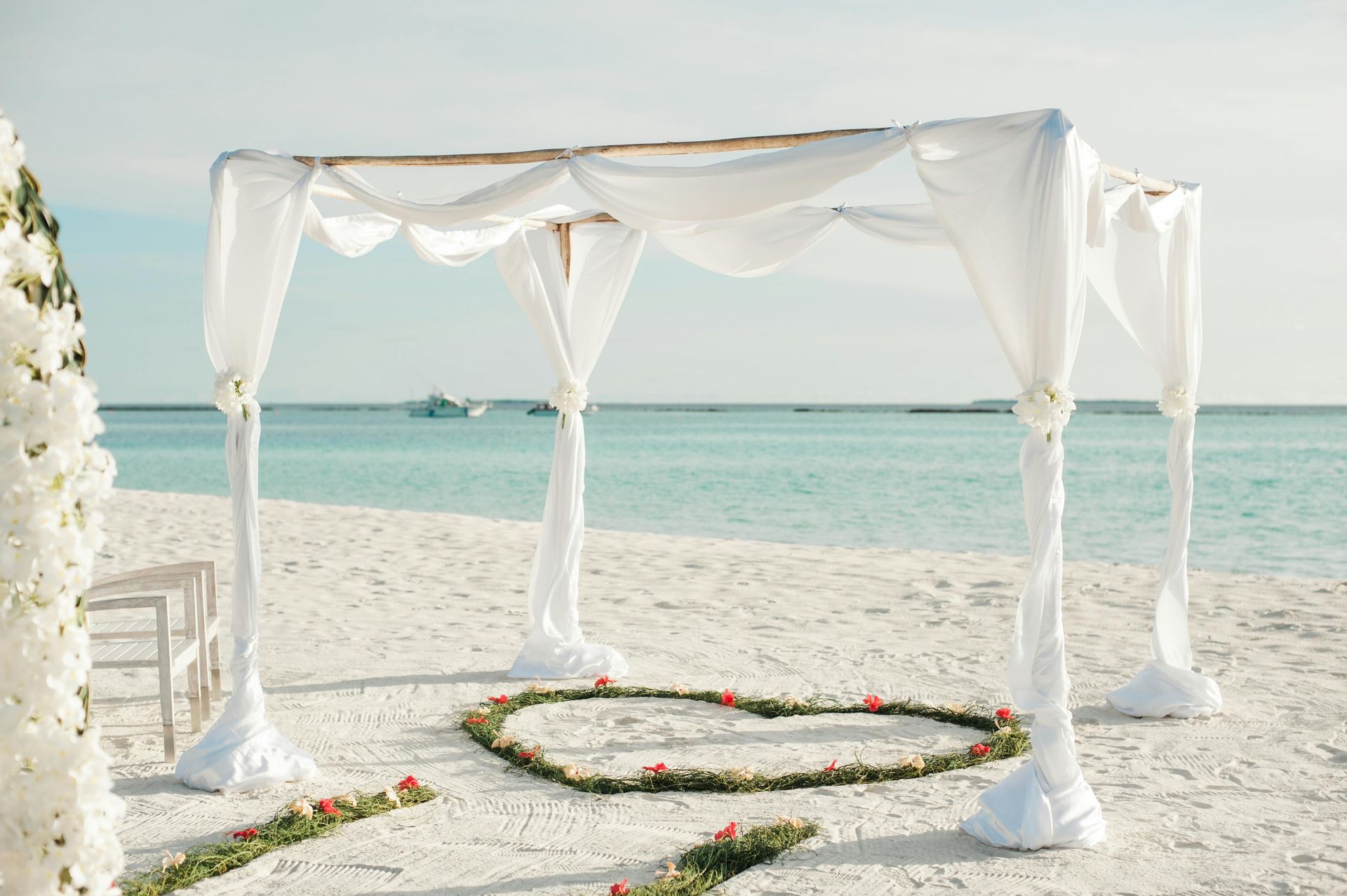 Beach wedding setup with white canopy, heart of flowers, ocean backdrop.