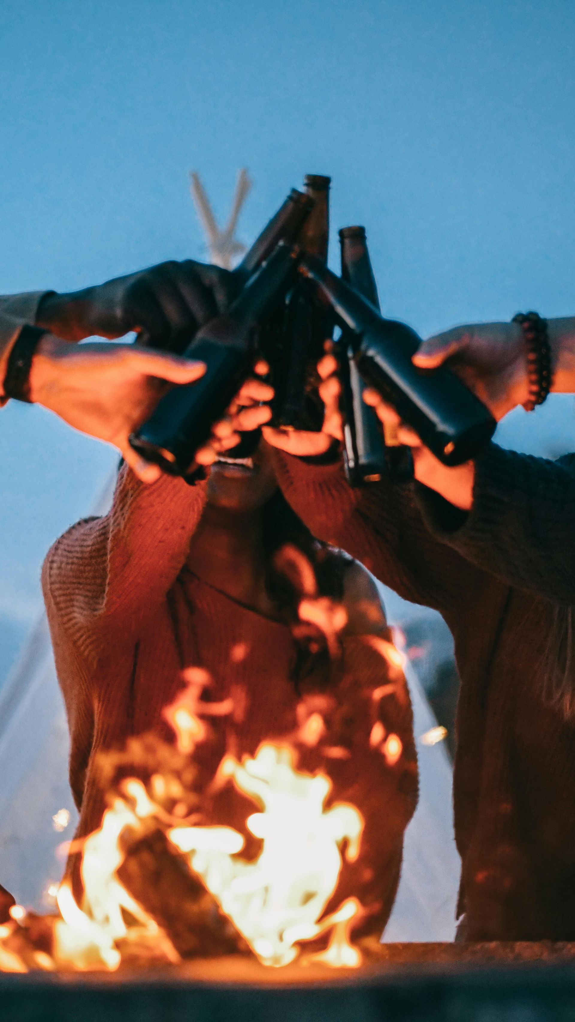Hands clinking dark bottles around a campfire, celebratory cheers.