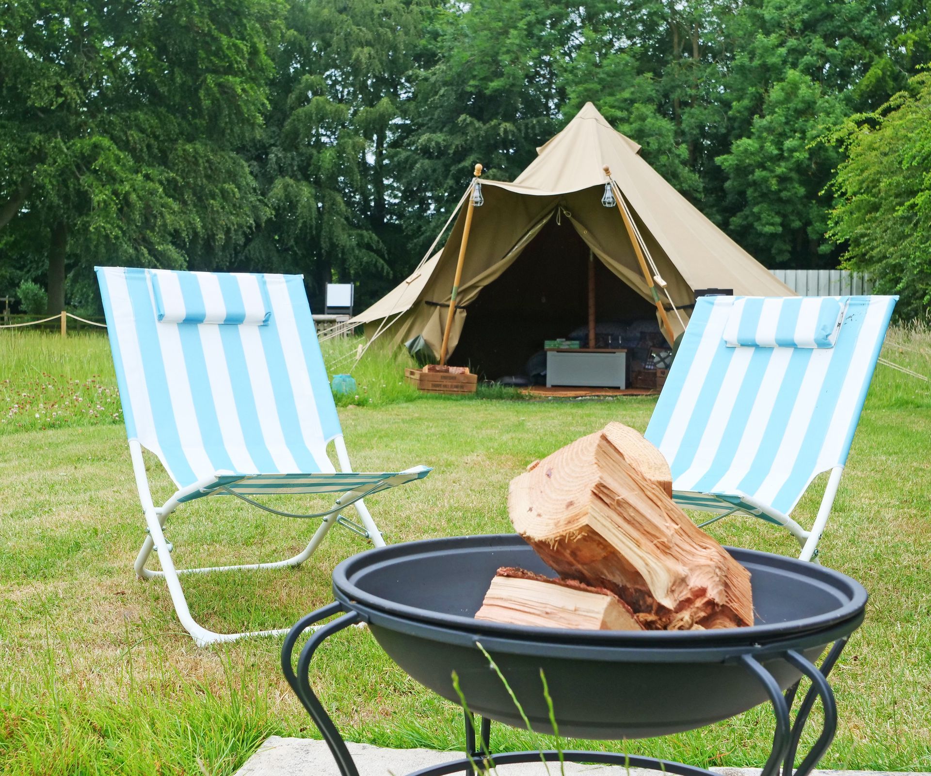 Two striped chairs and a fire pit in front of a tent in a grassy area, with trees in the background.