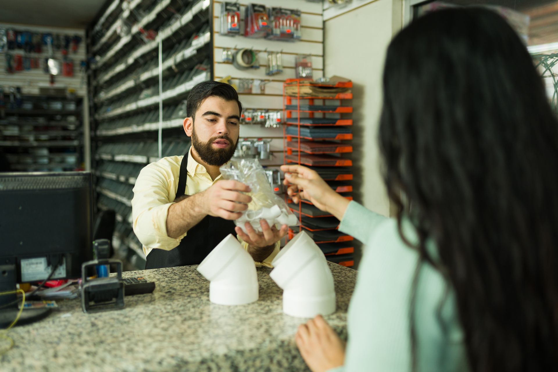 A store clerk hands a customer a bag over the counter, plumbing supplies visible. A store clerk hands a customer a bag over the counter, plumbing supplies visible.