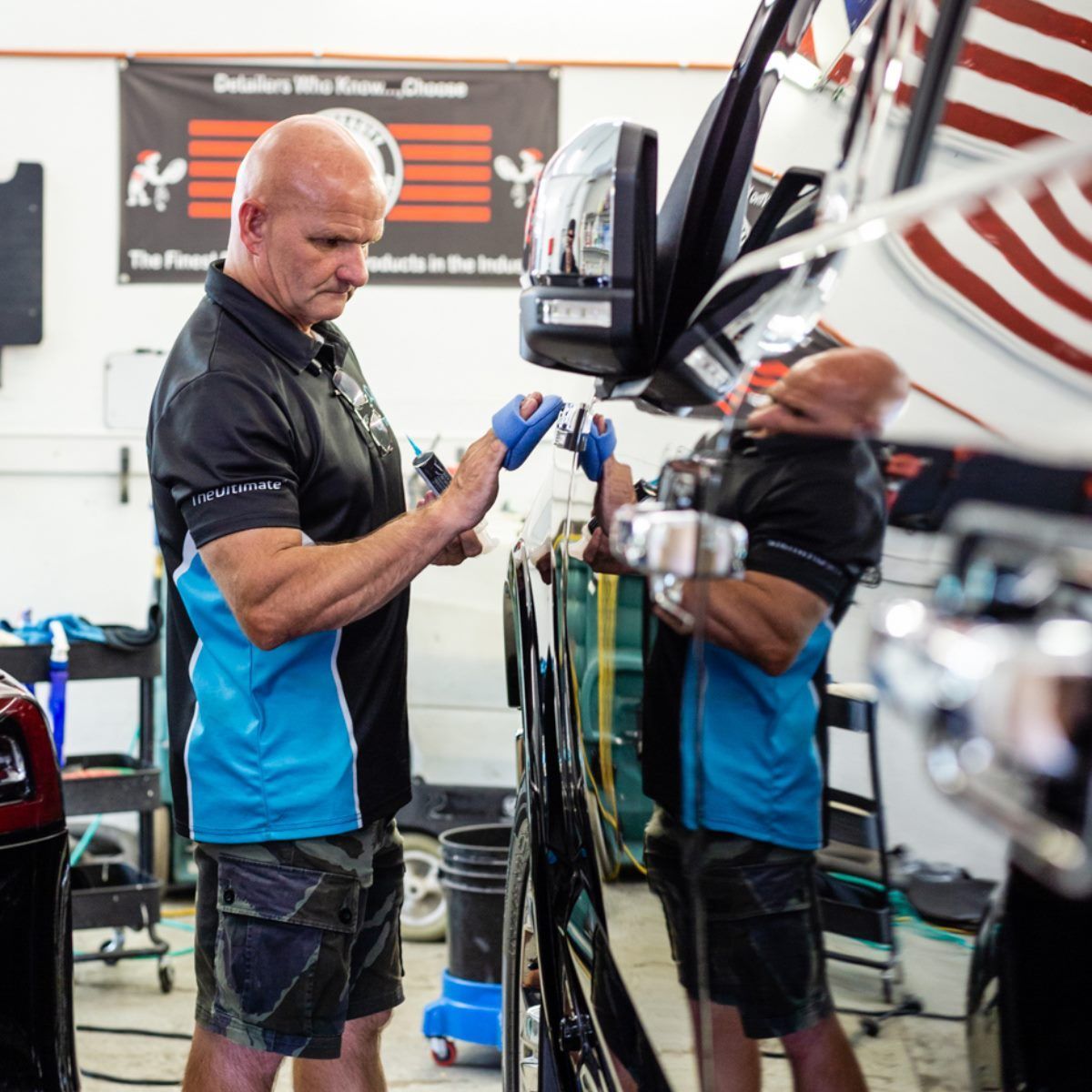 Bald man in shop, applying something to black car; blue gloves, black shirt, camo shorts.
