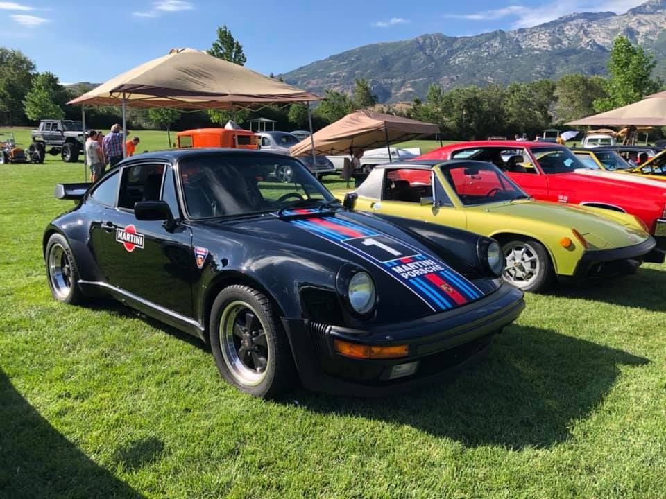 Black Porsche with racing stripes parked on grass at an outdoor car show, other classic cars in background.