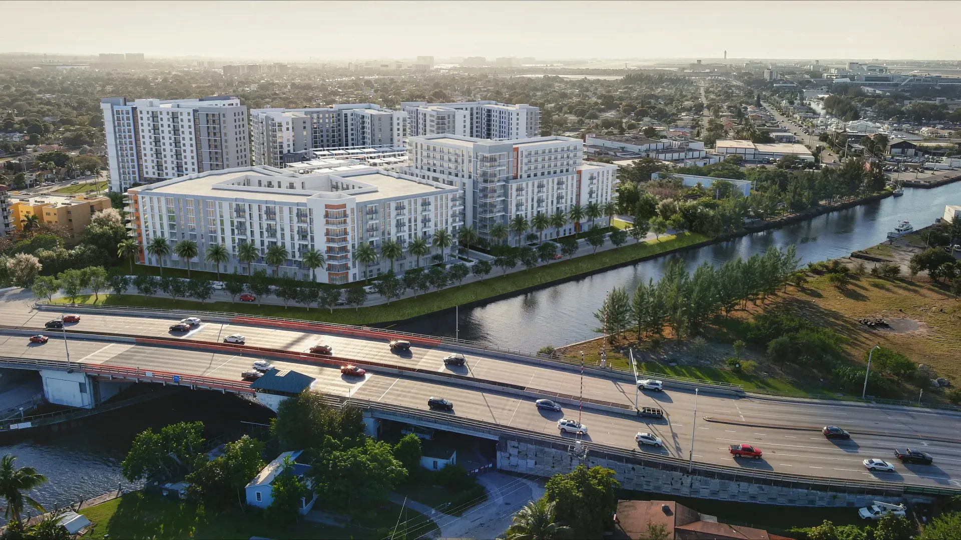 Aerial view of white apartment buildings beside a canal and bridge in a green urban neighborhood.