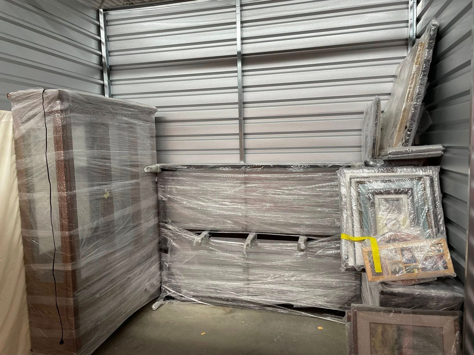 Wrapped furniture and framed items stacked in a warehouse against a corrugated metal wall