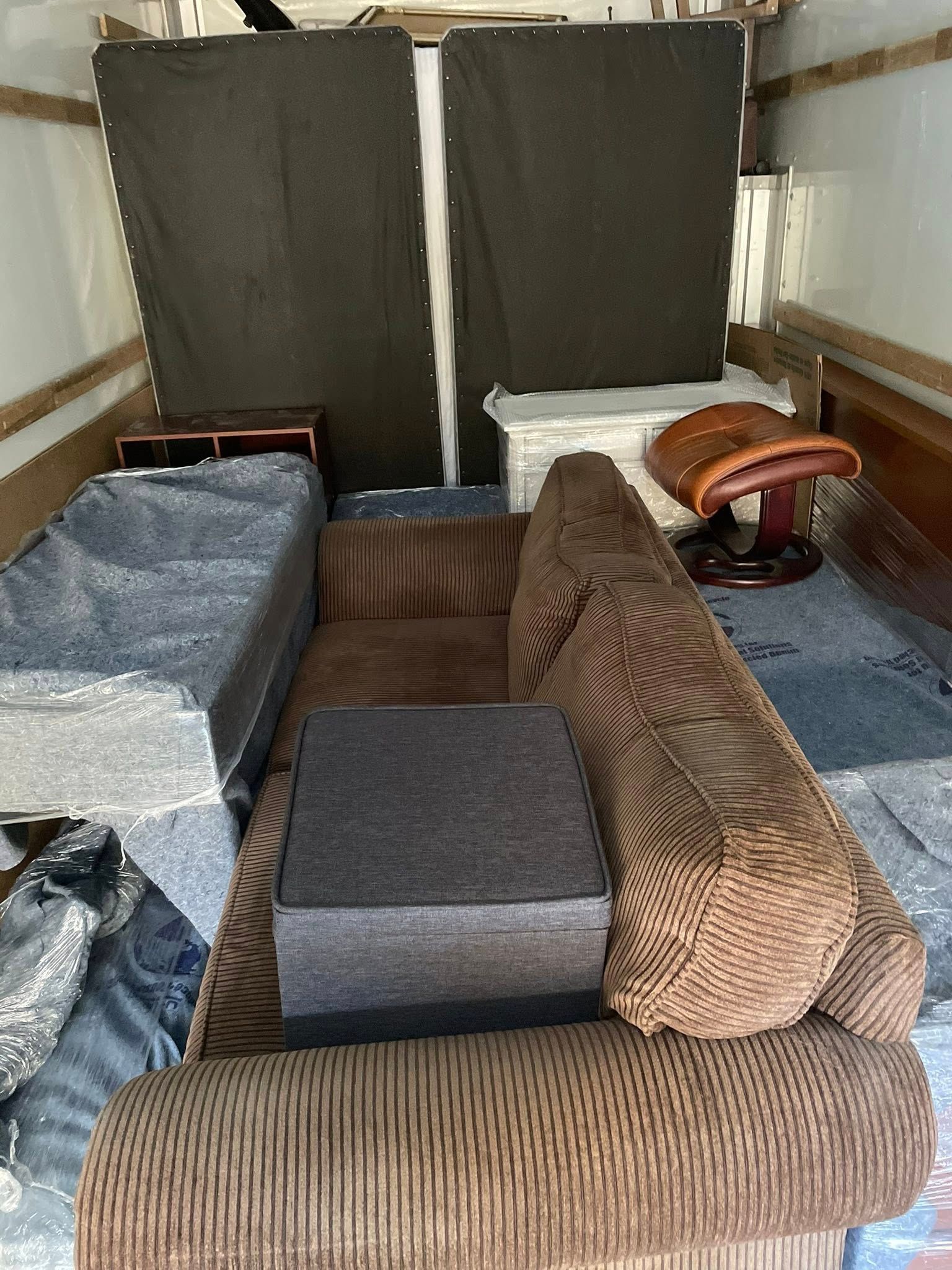 Truck cargo area filled with brown and gray sofas, cushions, and folded items, packed tightly against the back doors
