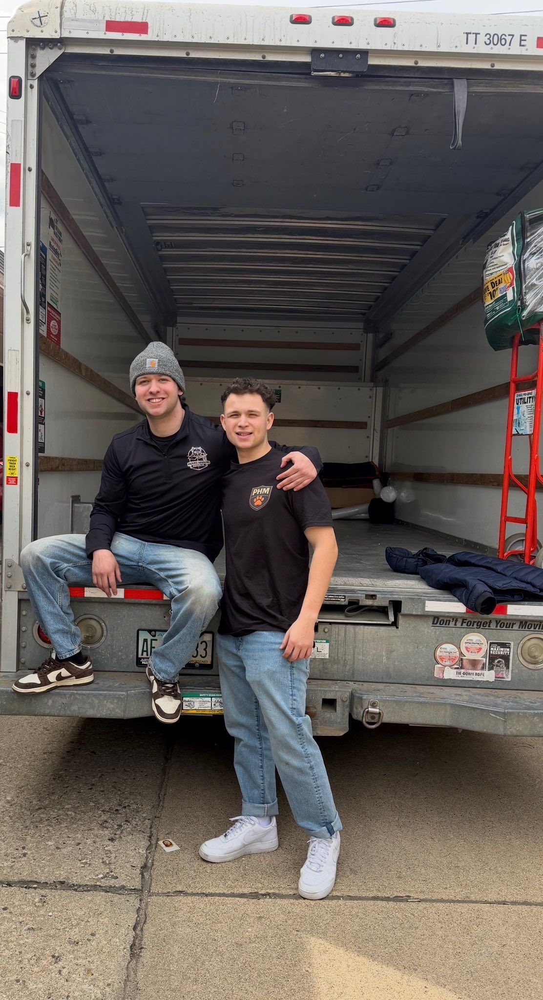 Two people posing at the back of an open box truck, one seated and one standing, with a ladder and gear inside.