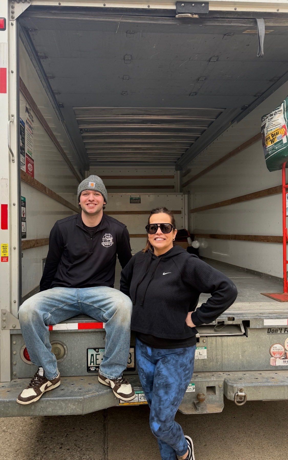 Two people posing in the back of an open moving truck, one seated and one standing, both in dark clothing.