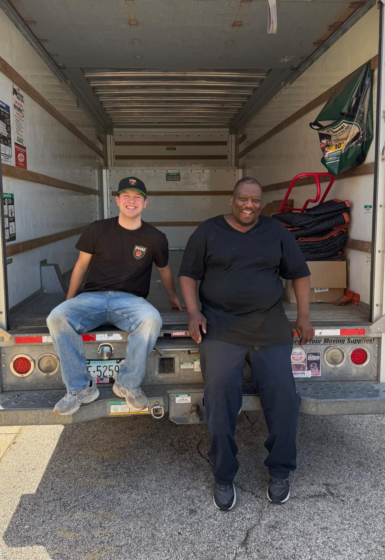 Two men sitting on the back of a moving truck, one in a cap and black shirt, in an open cargo area.
