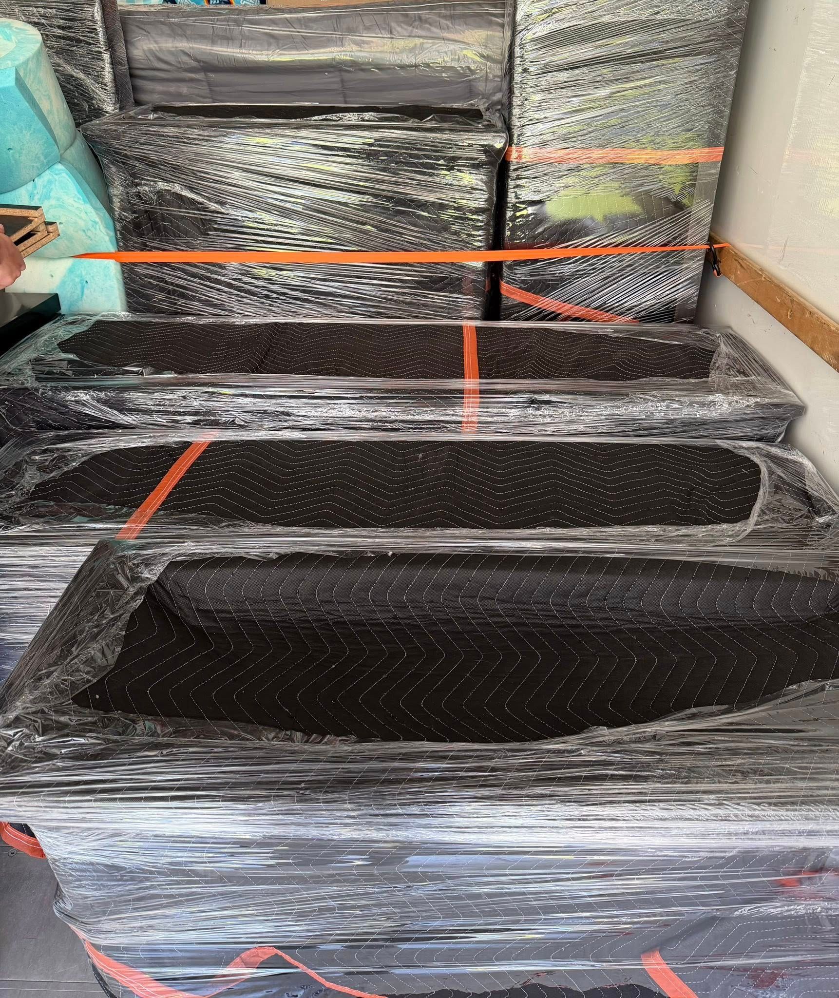 Stacked black furniture wrapped in plastic with orange straps, indoors against a wall.