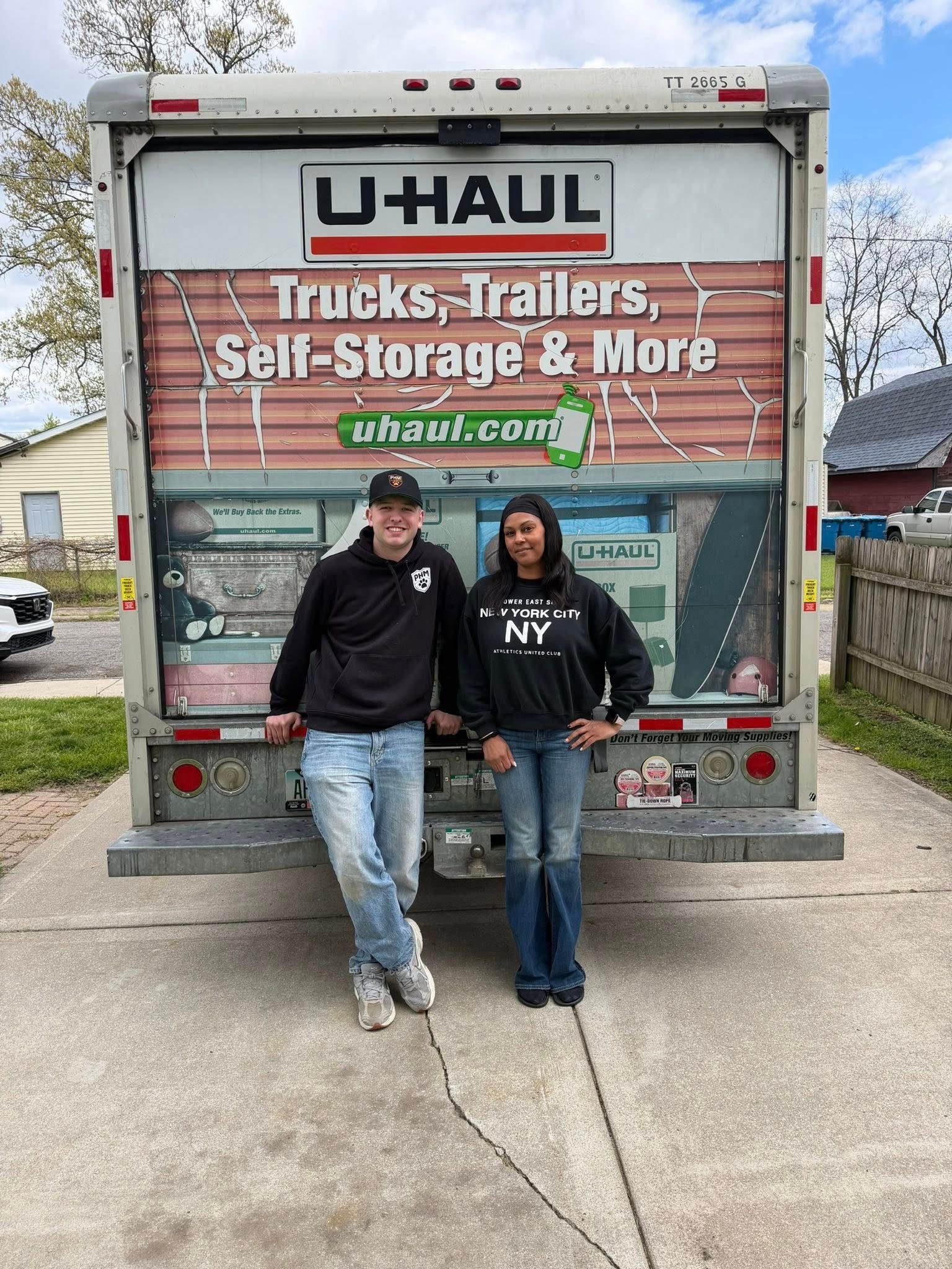 Two people standing in front of a U-Haul truck with a moving trailer sign.
