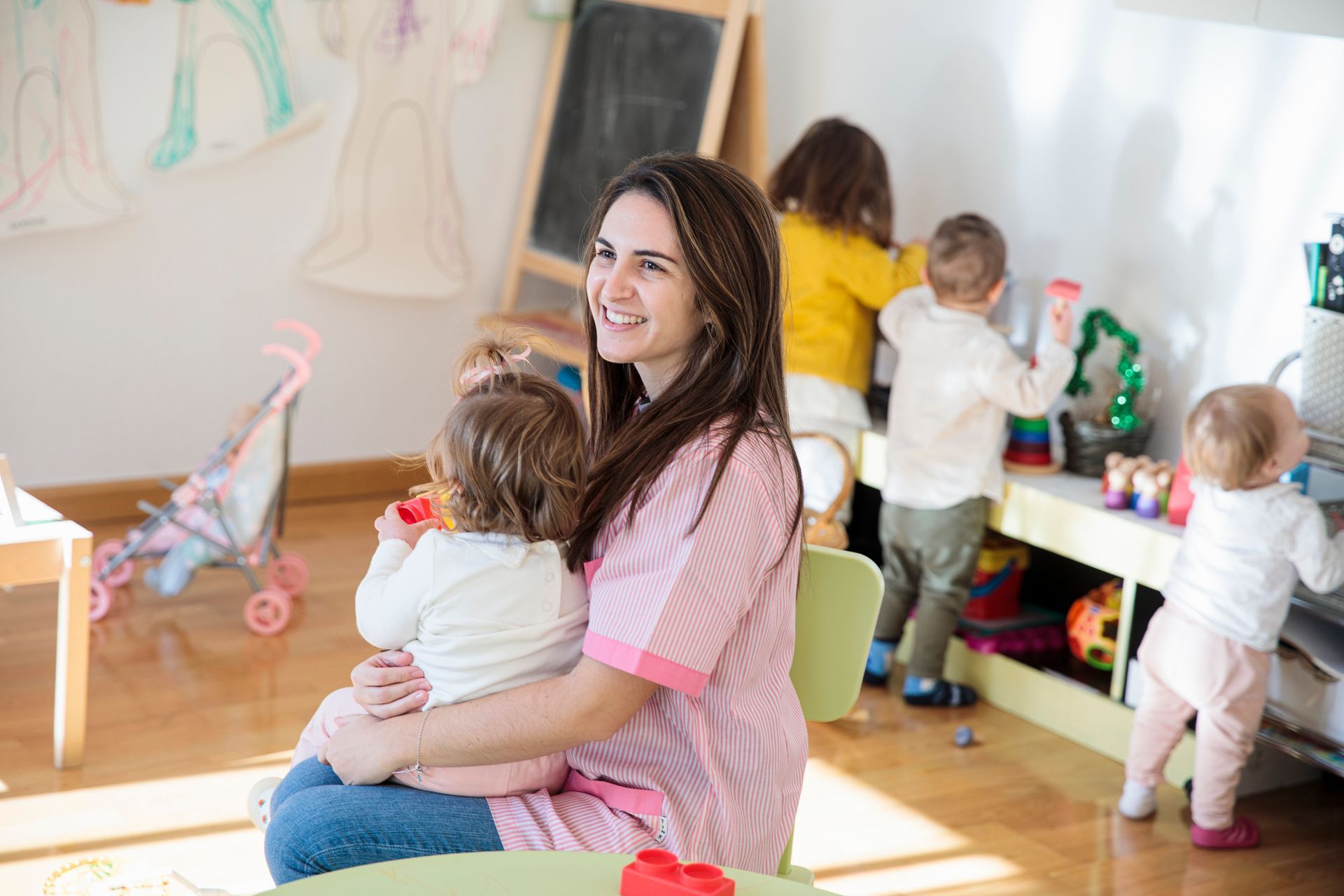 Female education teacher wearing pink t-shirt with a little girl in her arms at a daycare.