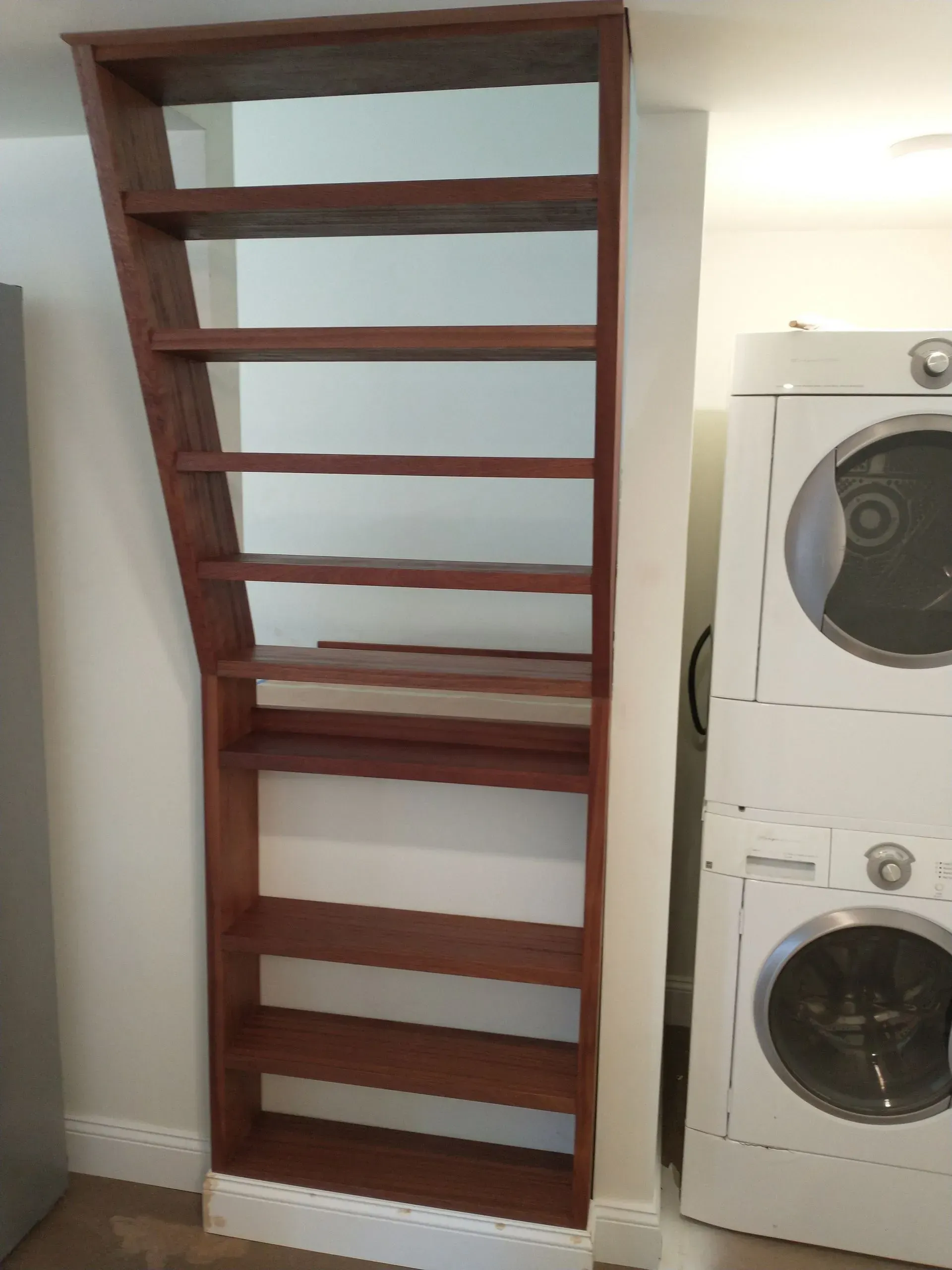 Tall, wooden, angled bookshelf built into a wall, next to stacked white washer and dryer.