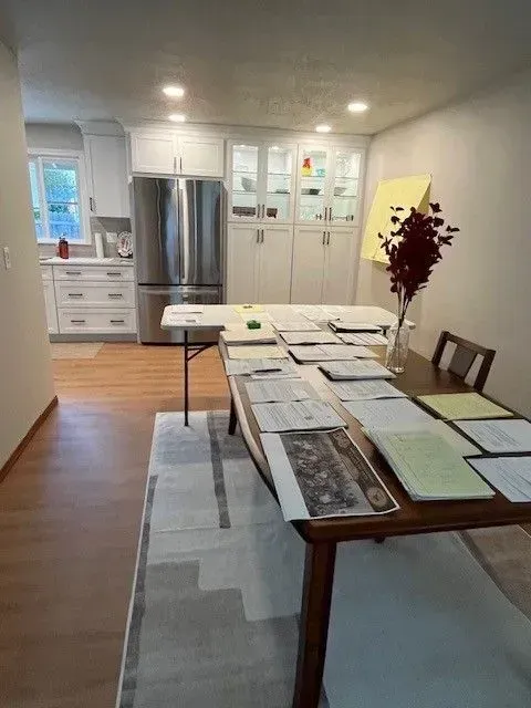 Kitchen and dining area with papers spread across a table and a folding table, cabinets, and a refrigerator.