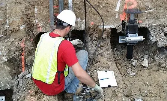 Construction worker examining a hole in the ground; wearing a hard hat and safety vest, working outdoors.