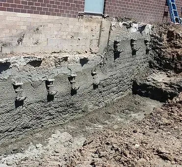 Foundation repair: concrete wall with steel anchors, exposed soil. Brick building in background.