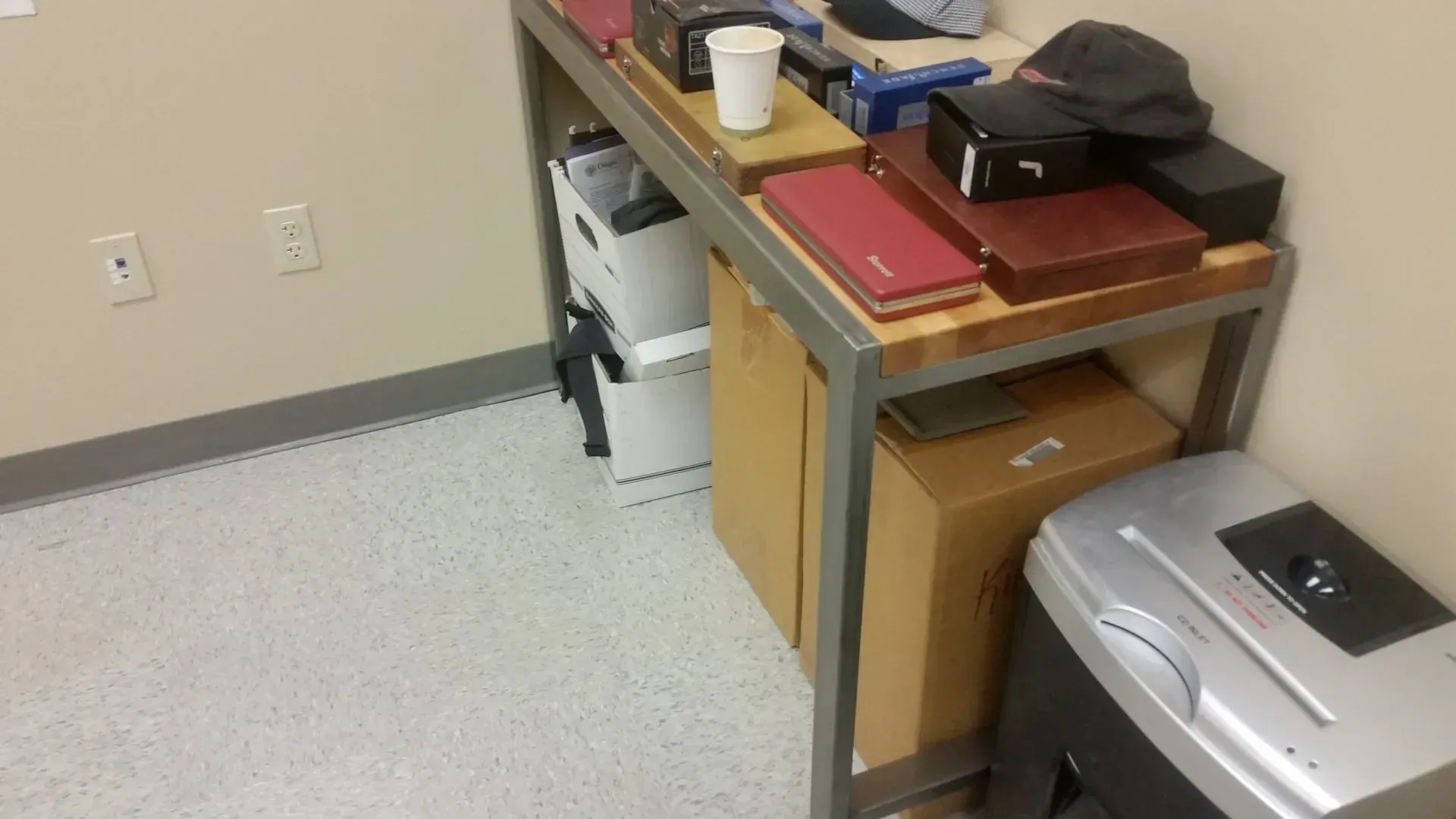 A desk with various office supplies, including boxes and a shredder, in the corner of a room.