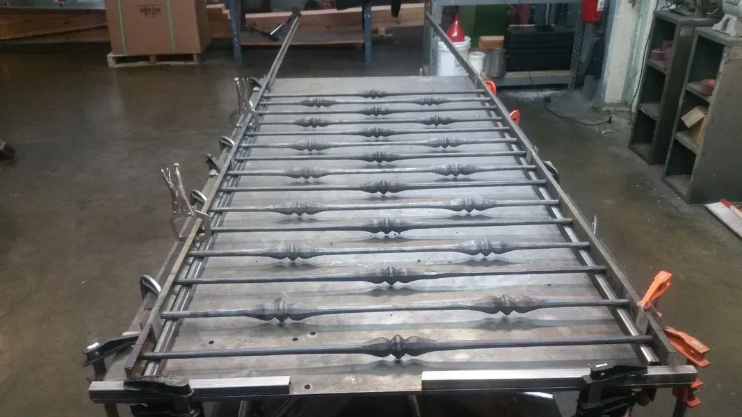 Metal frame with horizontal bars, some with decorative elements, clamped to a metal table in a workshop.