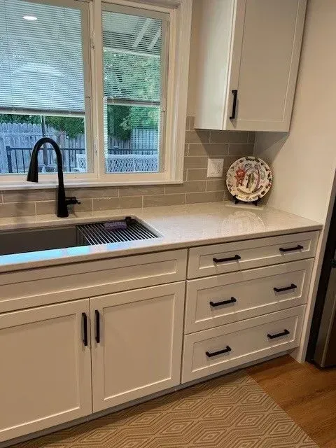 White kitchen cabinets with black handles, sink, window, and countertop, with a decorative plate.