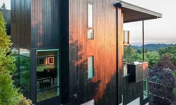 Modern multi-story house with dark wood siding, large windows, and a cantilevered balcony overlooking trees.