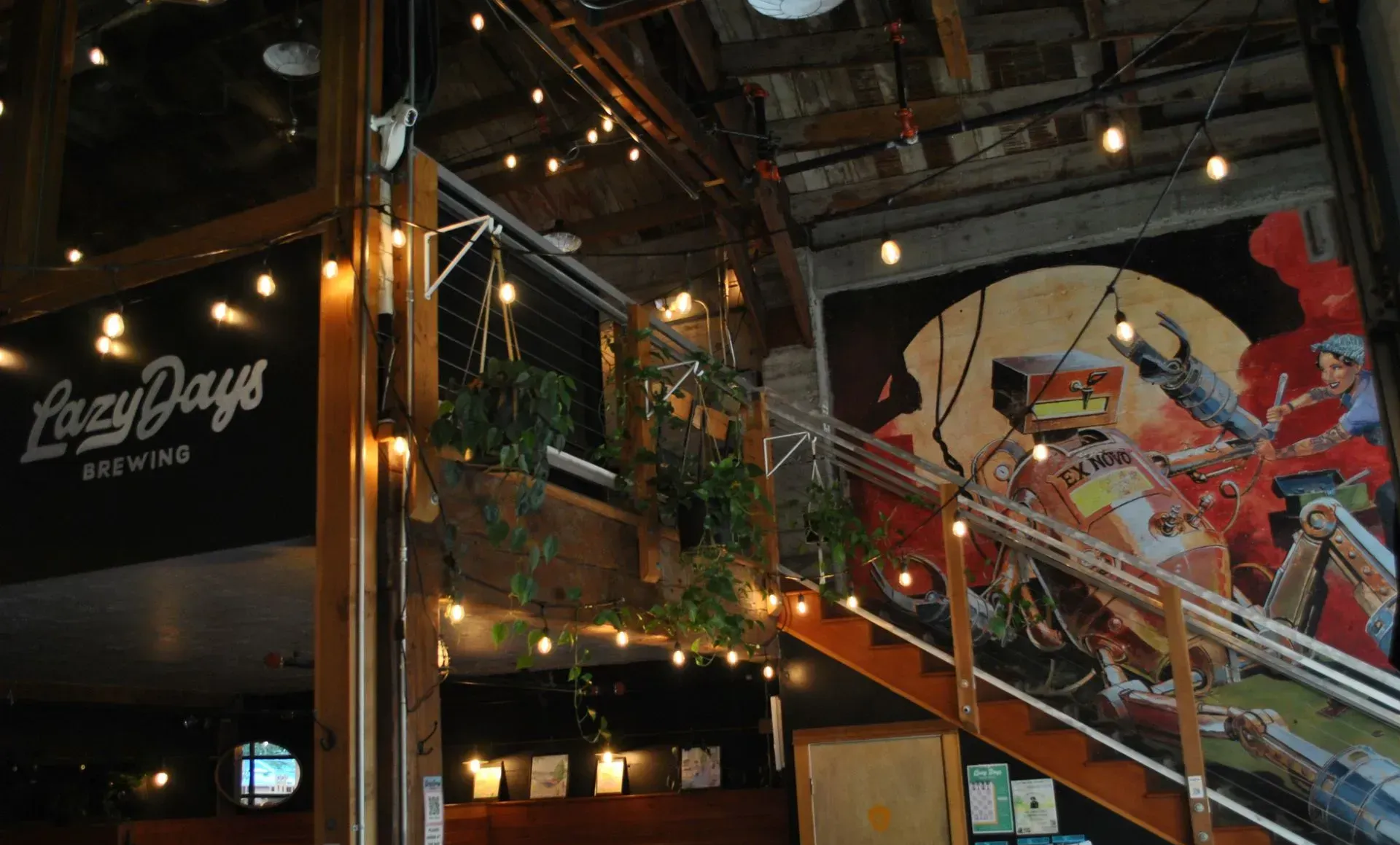 Interior of Lazy Days Brew Co. features a mural, string lights, wooden staircase, and hanging plants.