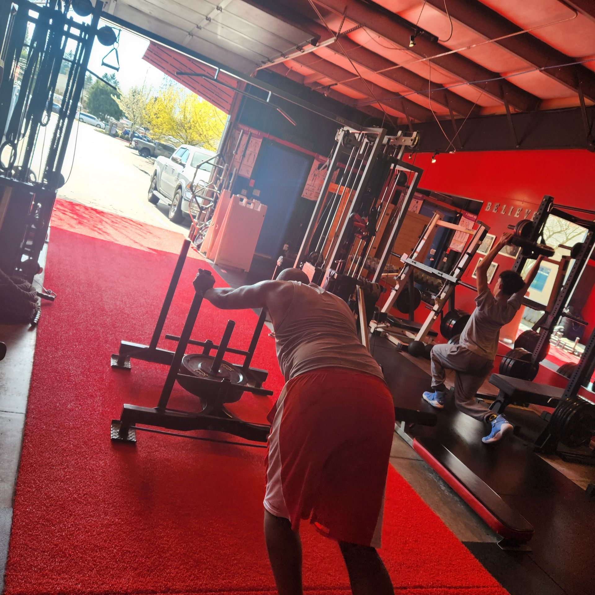 Two people exercising in a gym with red flooring; one pushes a weight sled while the other performs a cable exercise.