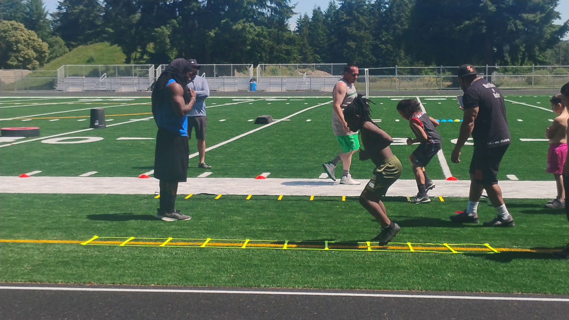 A group of people perform agility ladder drills on a grassy sports field under a bright, sunny sky.