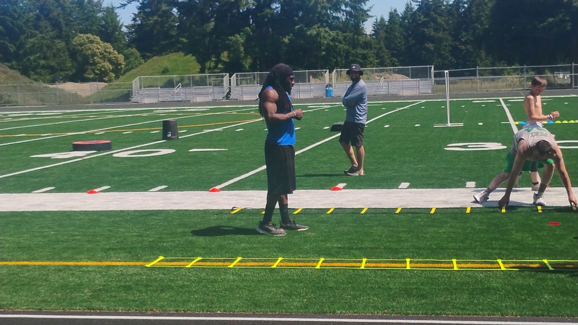 A coach observes athletes training with agility ladders on a sunny outdoor sports field.