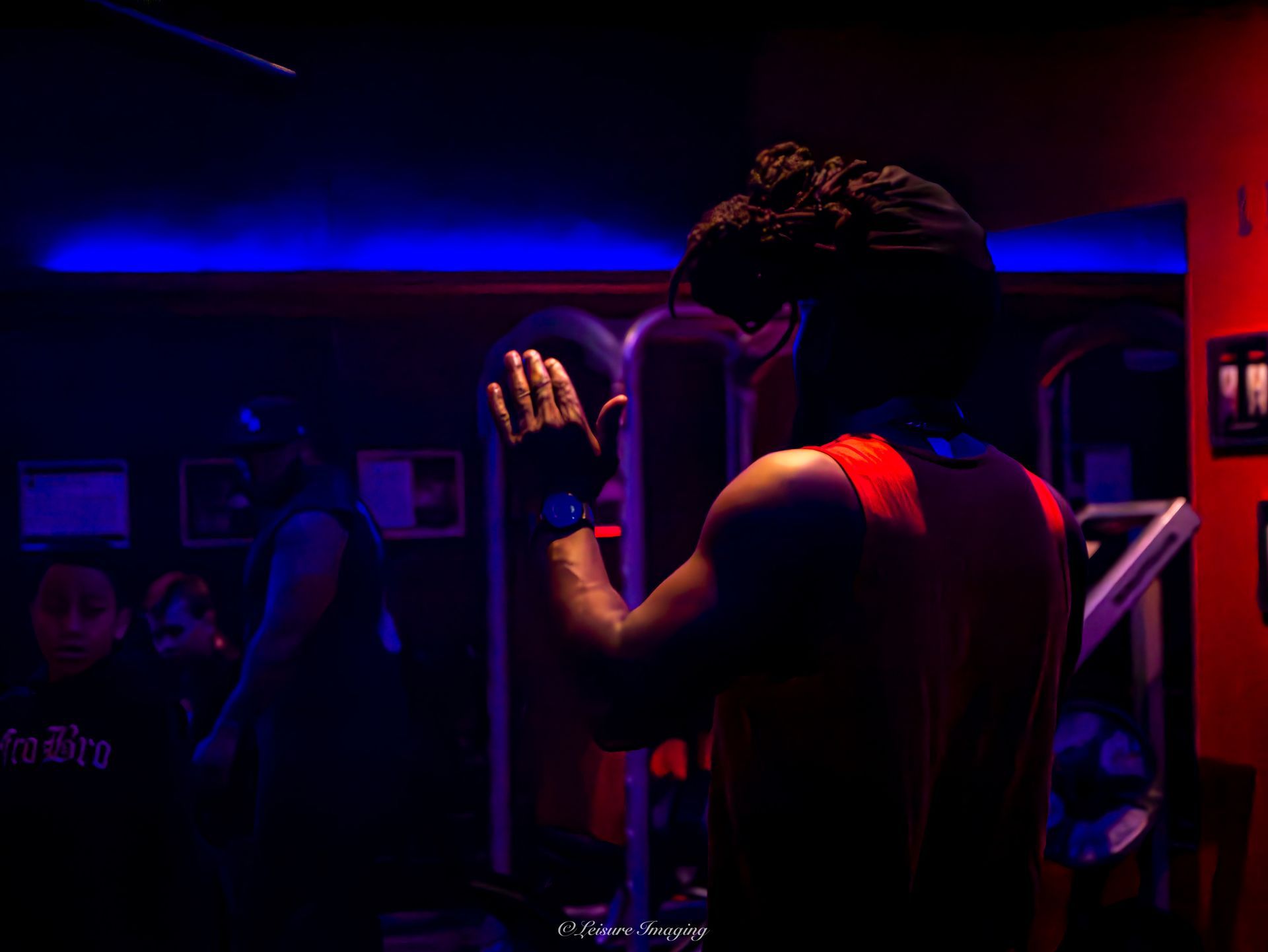 A person in a red tank top stands with their back to the camera, arm raised with an open palm in a dimly lit gym setting.