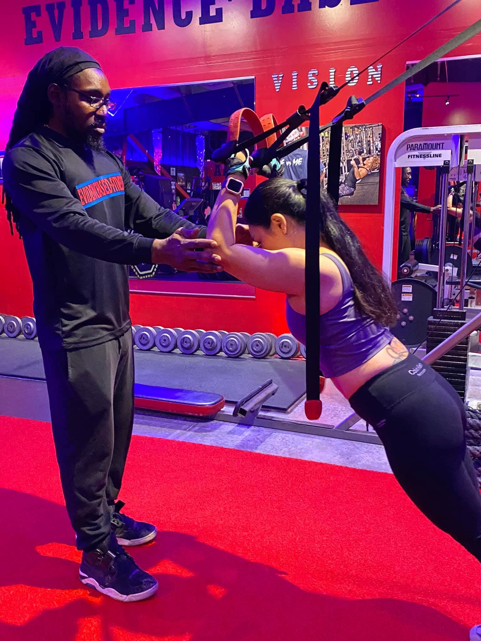 A trainer observes as a person performs a suspended plank exercise using suspension straps in a gym with red flooring.