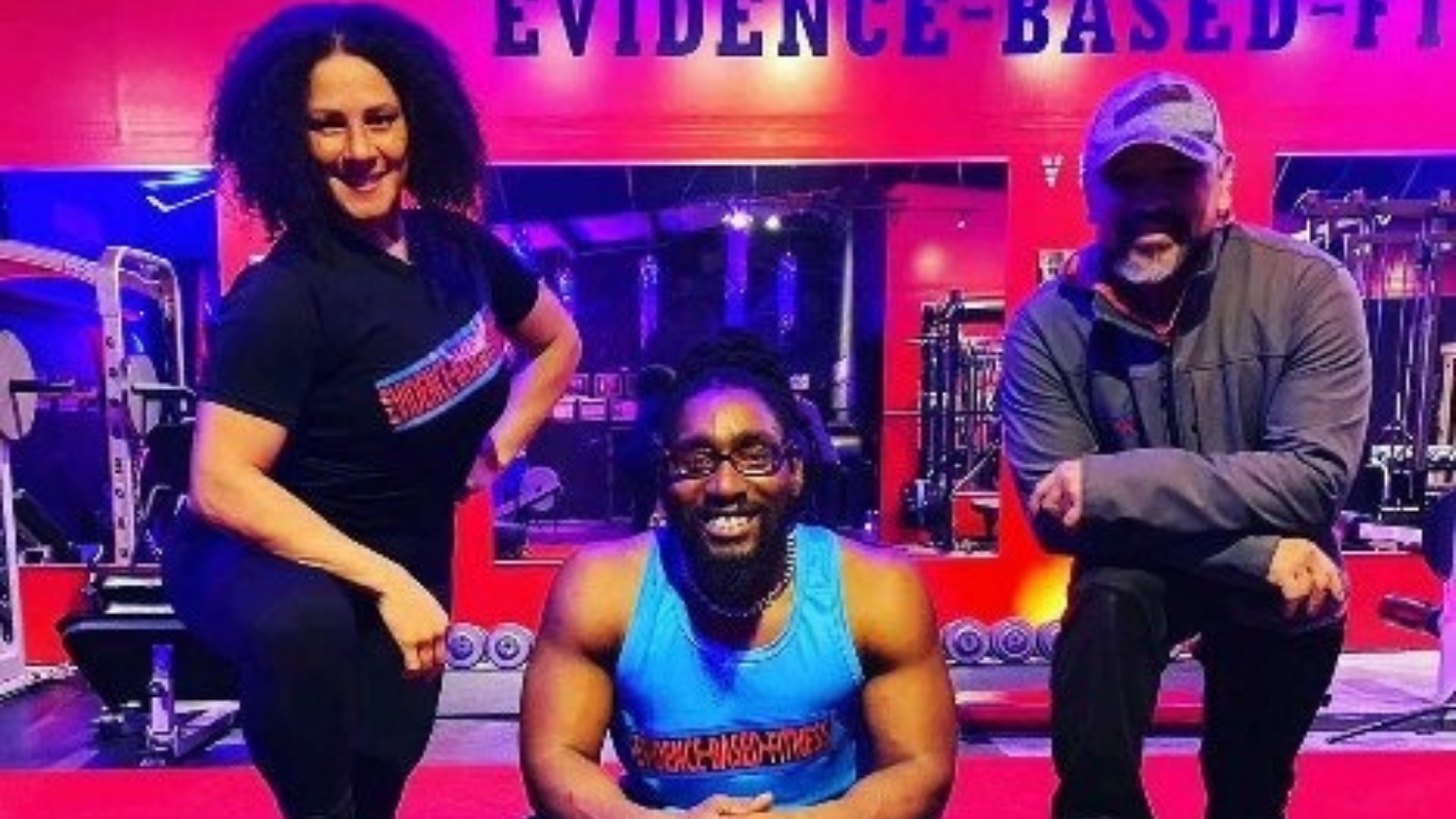 Three people smiling in a gym, posing near exercise equipment. Sign in background reads 