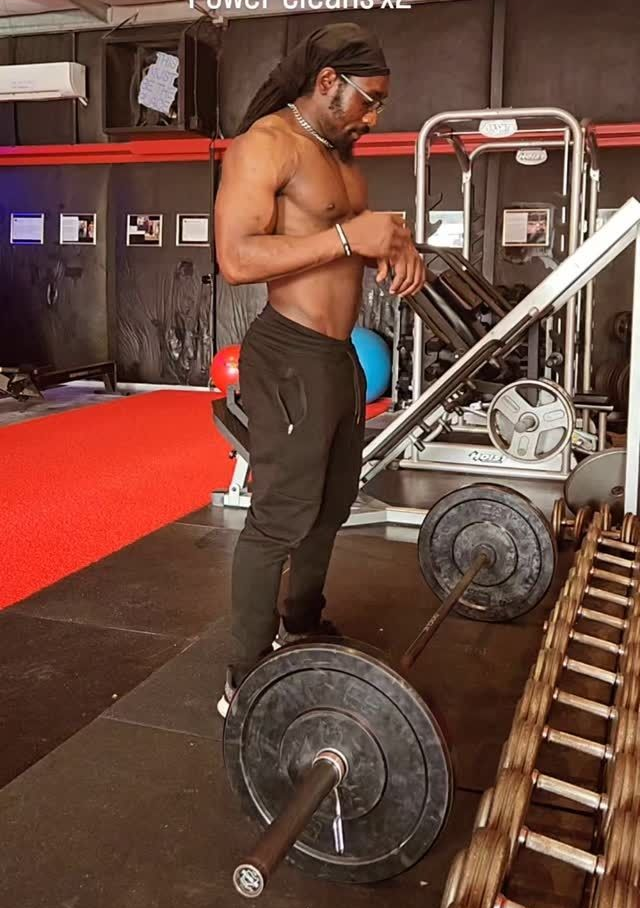 Man in gym preparing to lift a barbell. Black pants, dreadlocks, black gym. Weights are on the floor.