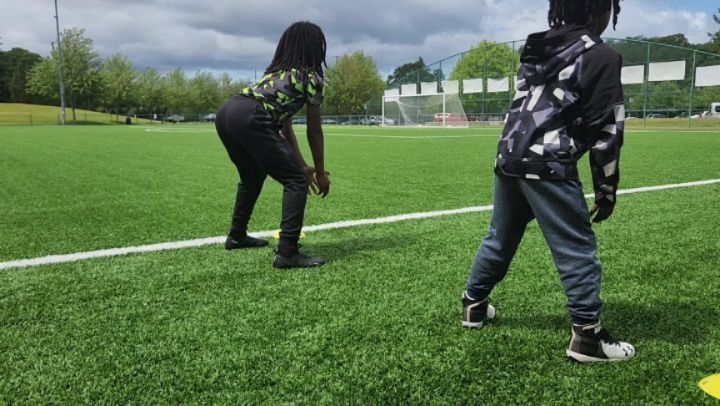 Two people exercising on a green field. One wears black and patterned shirt, the other a grey hoodie.