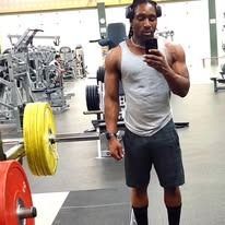 Man in gym, lifting weights. Wearing headphones, grey tank, black shorts.