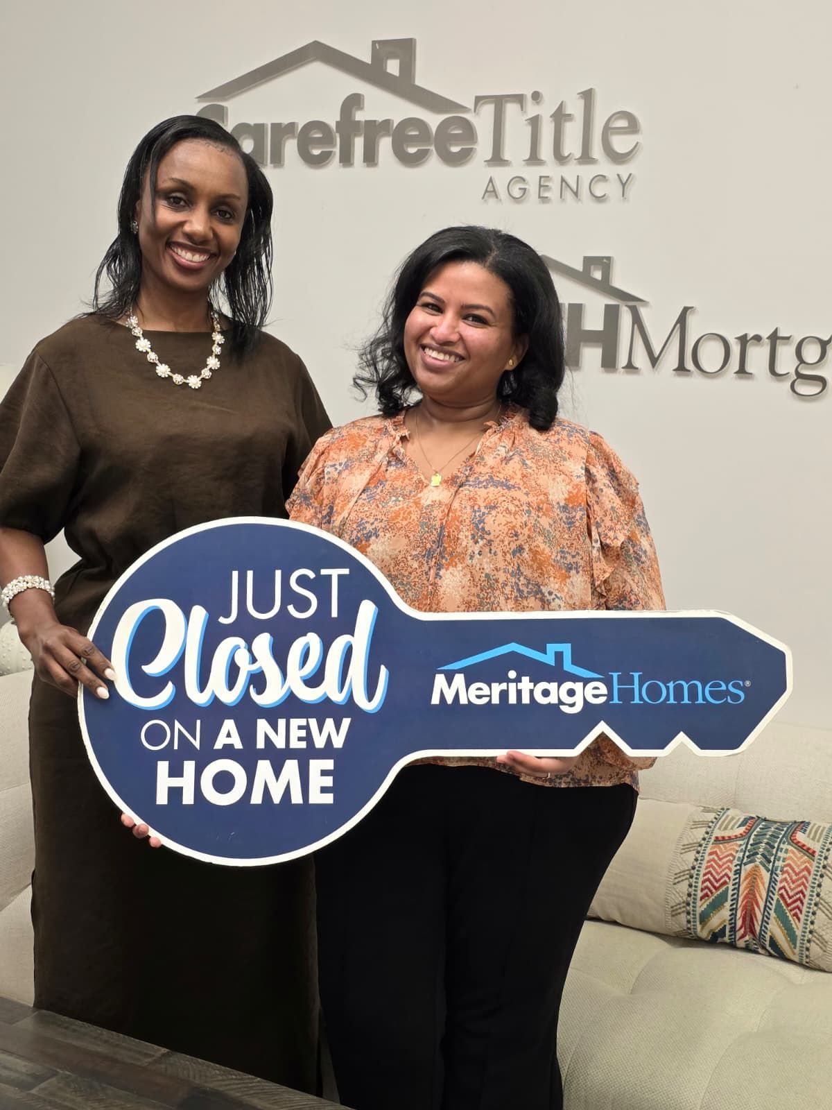 Two women holding “Just Closed on a New Home” and Heritage Homes signs in front of a realty backdrop