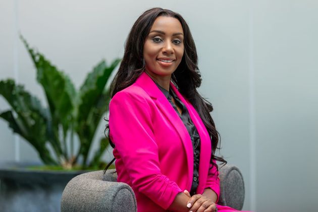 Woman in a bright pink blazer seated indoors, smiling beside green plants.