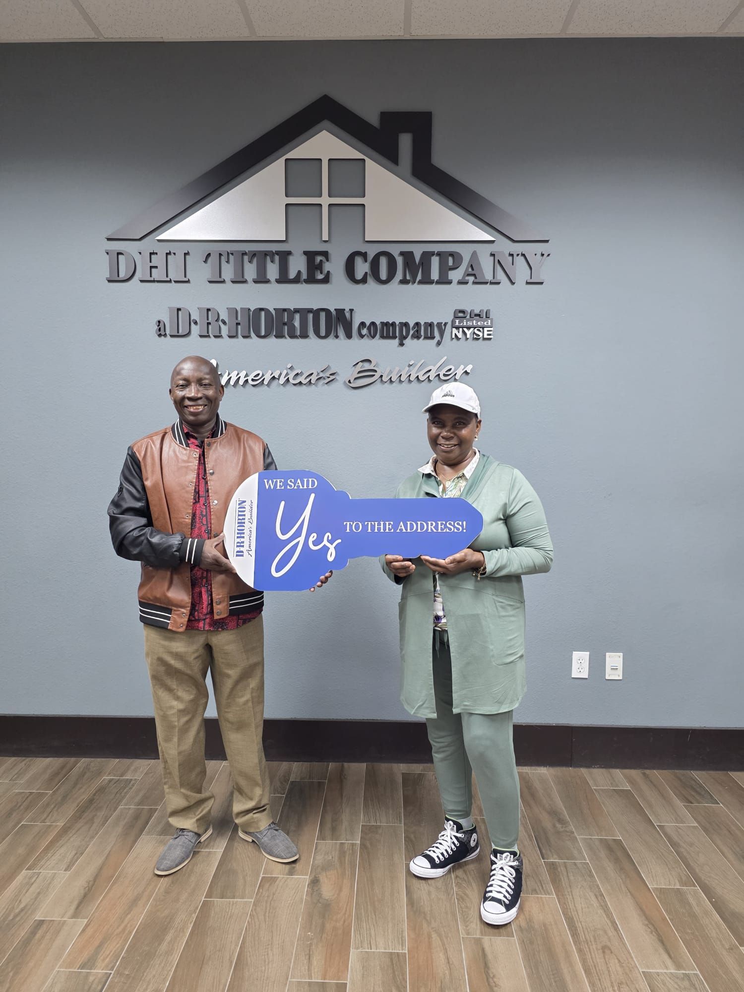 Two people holding a blue key-shaped sign in front of a DHI Title Company wall logo.