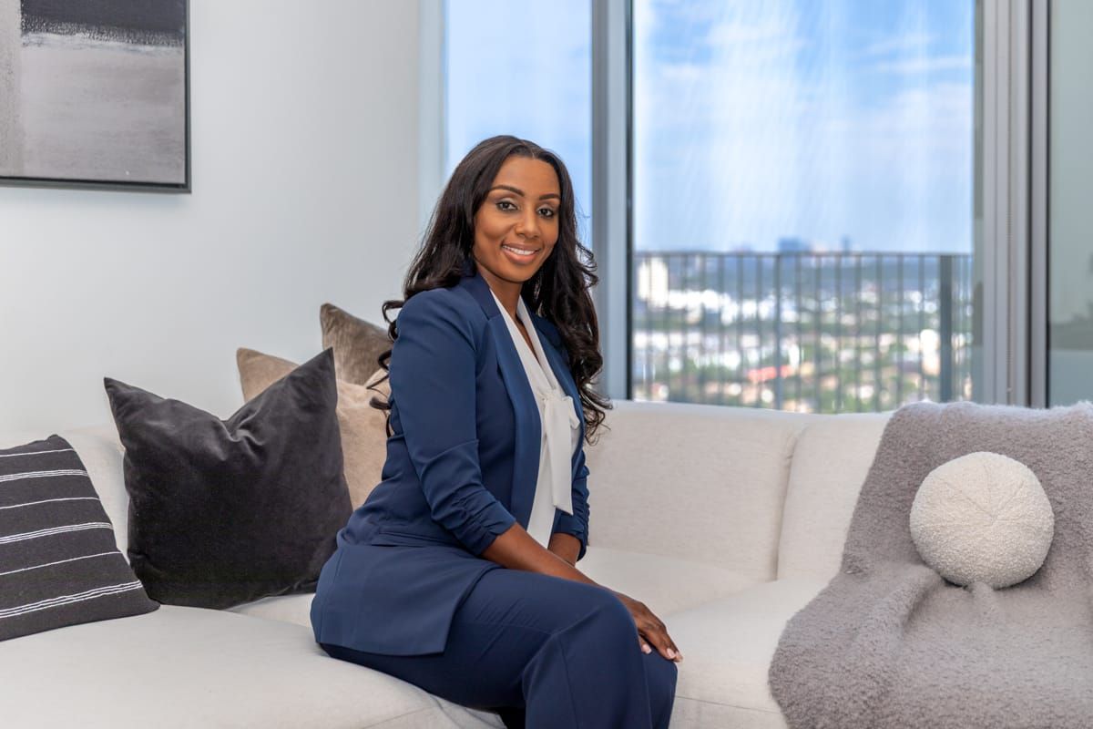 Woman in a navy suit seated on a light sofa in a modern living room, smiling at the camera.