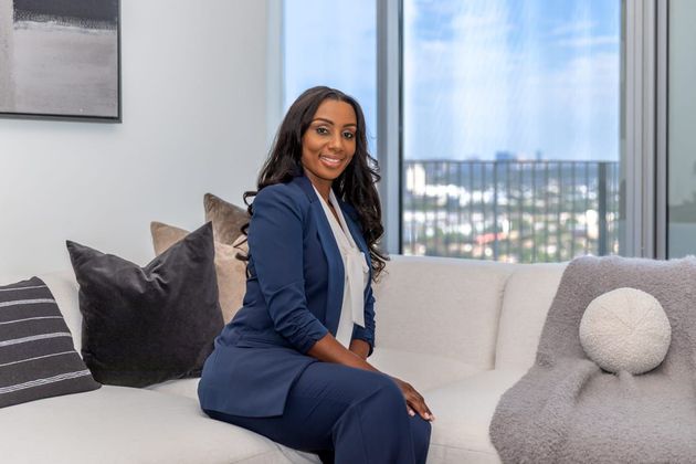 Woman in a navy suit seated on a light sofa in a modern living room, smiling at the camera.